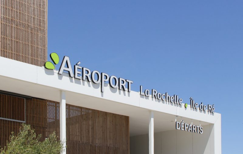 Photographie du haut du auvent de l'aéroport avec le nom de l'aéroport en signalétique blanc sur fond de ciel bleu
