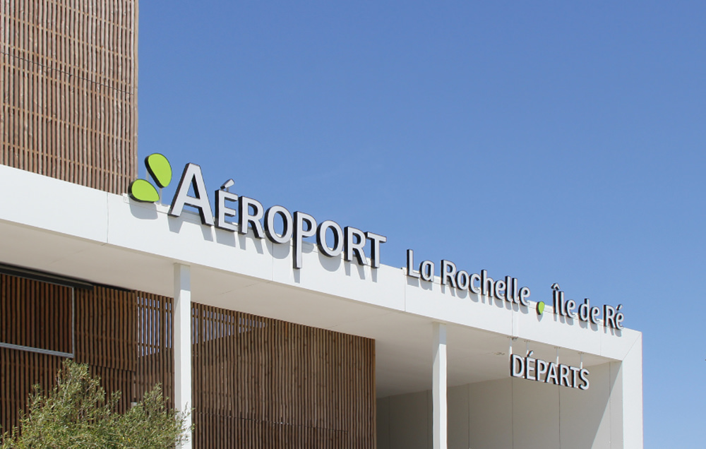 Photographie du haut du auvent de l'aéroport avec le nom de l'aéroport en signalétique blanc sur fond de ciel bleu