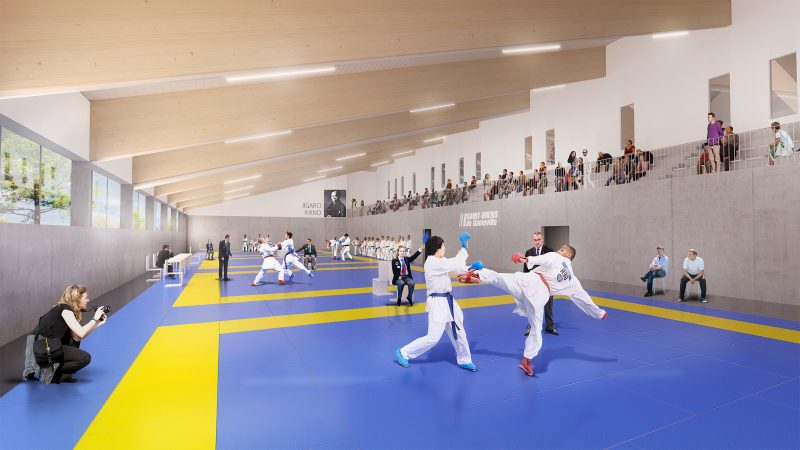 Perspective d'intérieur de la salle de judo, les spectateurs dans les gradins en hauteurs observent une compétition, de l'autre côté des gradins une baie vitrée sur toute la longeur éclaire la scéne
