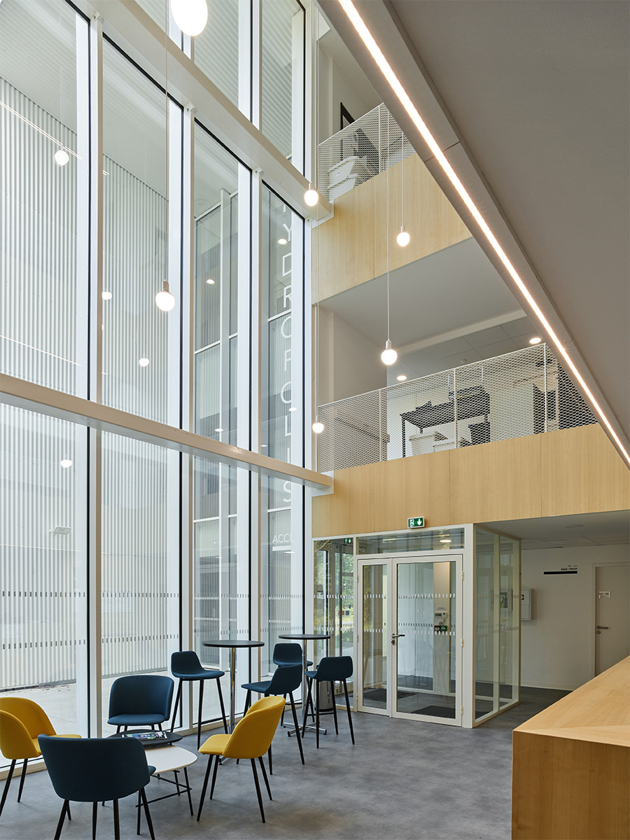 Photographie depuis l'accueil sous la mezzanine, vue sur les deux etages au dessus et sur le sas d'entrée, tables et fauteuils à disposition