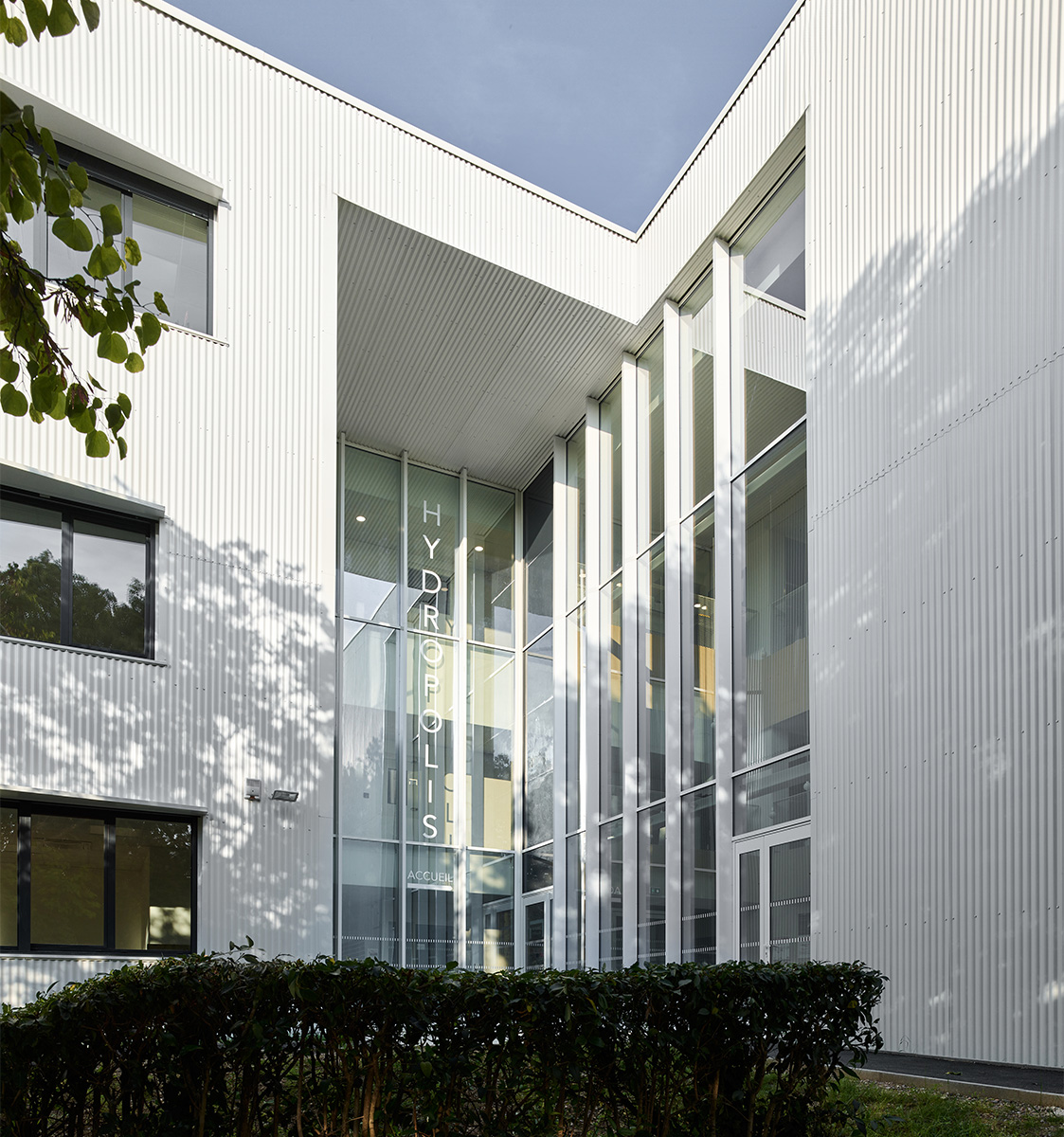 Photographie de l'entrée de Hydropolis à Montpellier, dans la cours entre les deux façades au bardage métallique vertical blanc, l'entrée vitrée sur deux niveaux apporte de la lumiére