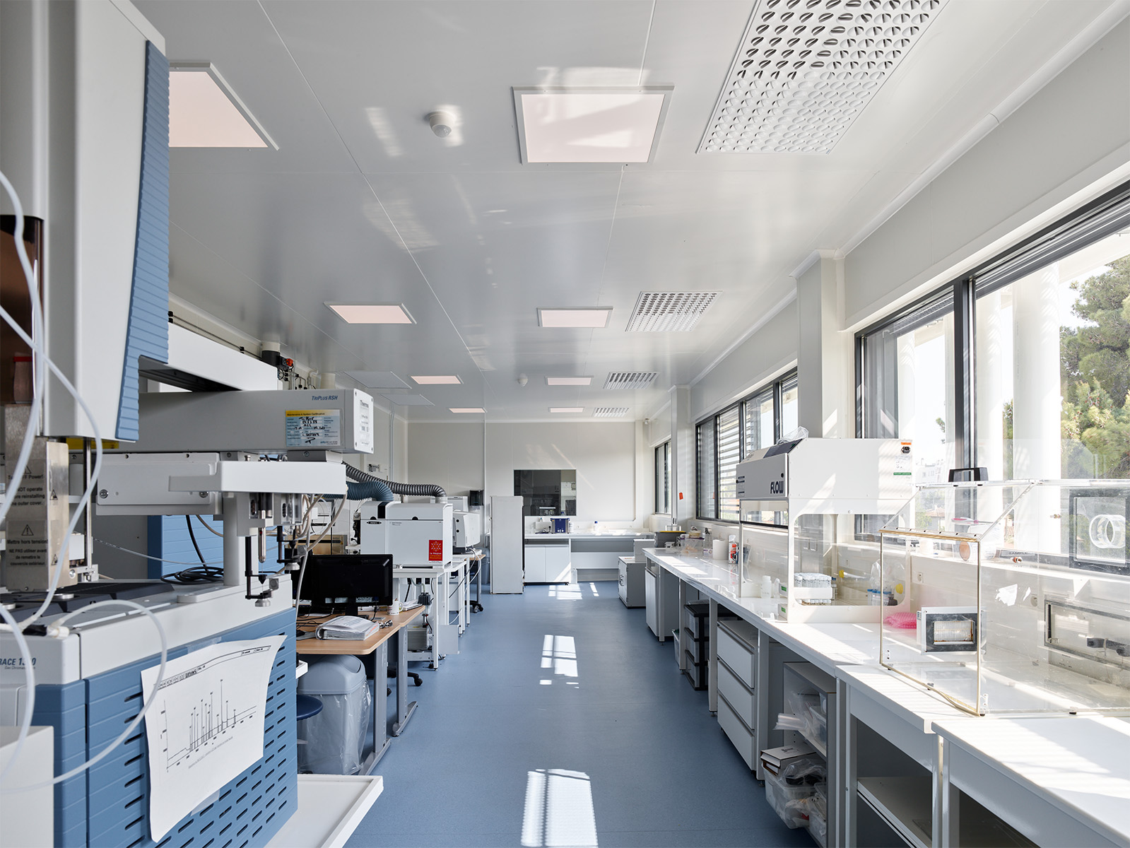 Photographie d'un laboratoire avec divers machines et tuyaux, sol bleu gris et mur et plafond blanc, les fenêtres en bandeaux apportent beaucoup de lumière