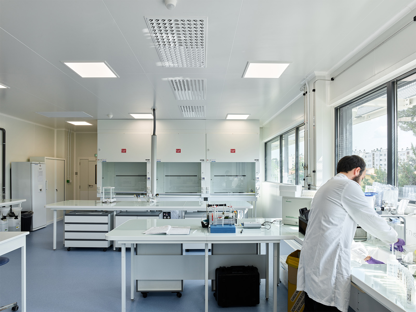 Photographie d'un laboratoire avec une personne travaillant en blouse blanche, sol bleu gris et mur et plafond blanc, les fenêtres en bandeaux apportent beaucoup de lumière