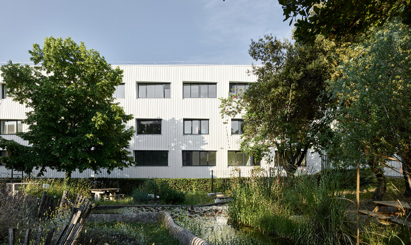 Photographie depuis la cour intérieure d'Hydropolis avec un bassin végétalisé entouré de grands arbres, en arrière plan le bâtiment blanc sur trois niveaux menuiserie gris anthracite
