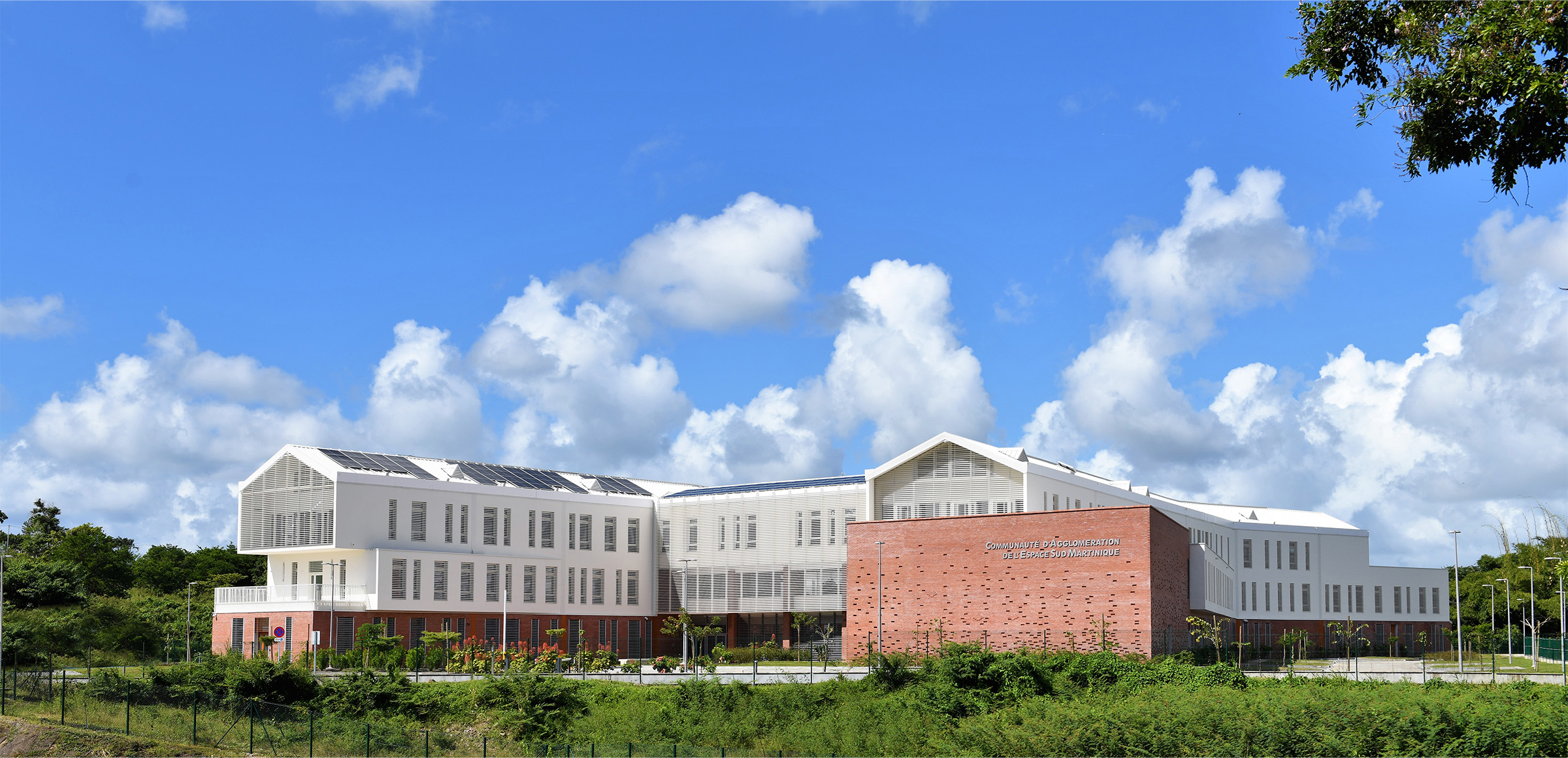 Photographie d'ensemble du bâtiment en plein jour depuis l'espace végétal, vue sur le bâtiment blanc en forme de H, soubassement brique et contexte végétal sauvage