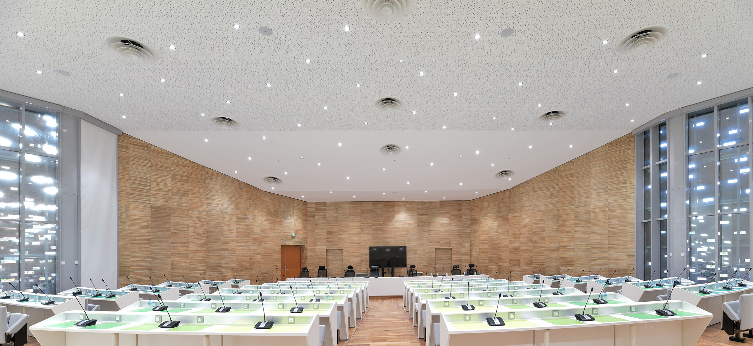 Photographie depuis le haut de la salle de conférence, teinte bois au sol et façade, baies vitrées vers l'entrée de la salle ne laissant passer qu'un filet de lumiére rappelant le jeu de lumiére au plafond comme un ciel étoilé, les tables dans l'amphithéâtre sont blanches avec un dégradé du jaune au bleu à l'extrémité de la salle