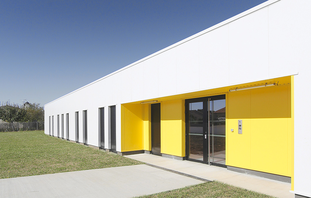 Photographie de trois quarts du bâtiment blanc sur 1 étage, entrée avec un cadre jaune