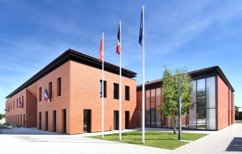 Photographie vue de trois quarts de la mairie vêtue de briques, trois drapeaux flottent dans l'espace vert