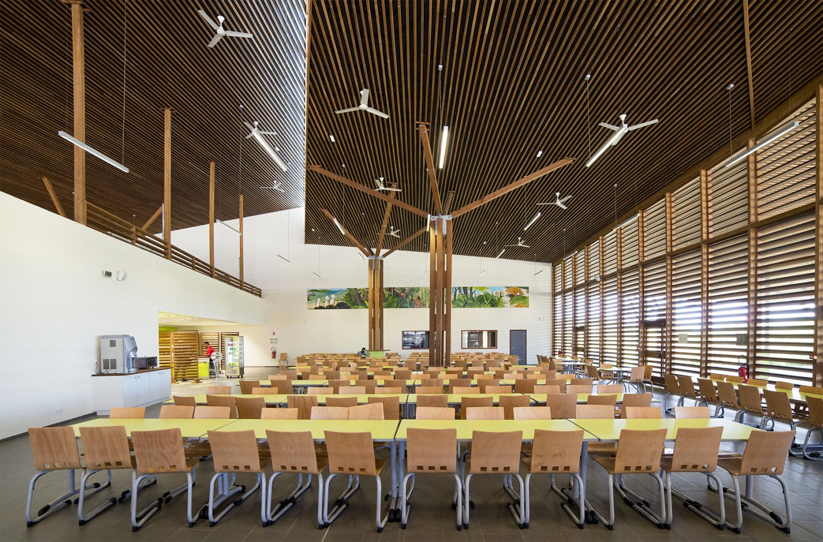 Photographie face aux places assises rangées du self, mur blanc et ouverture sur l'exterieur tamisée par des lames bois horizontales tout hauteur, plafond en lames de bois et ventilateurs