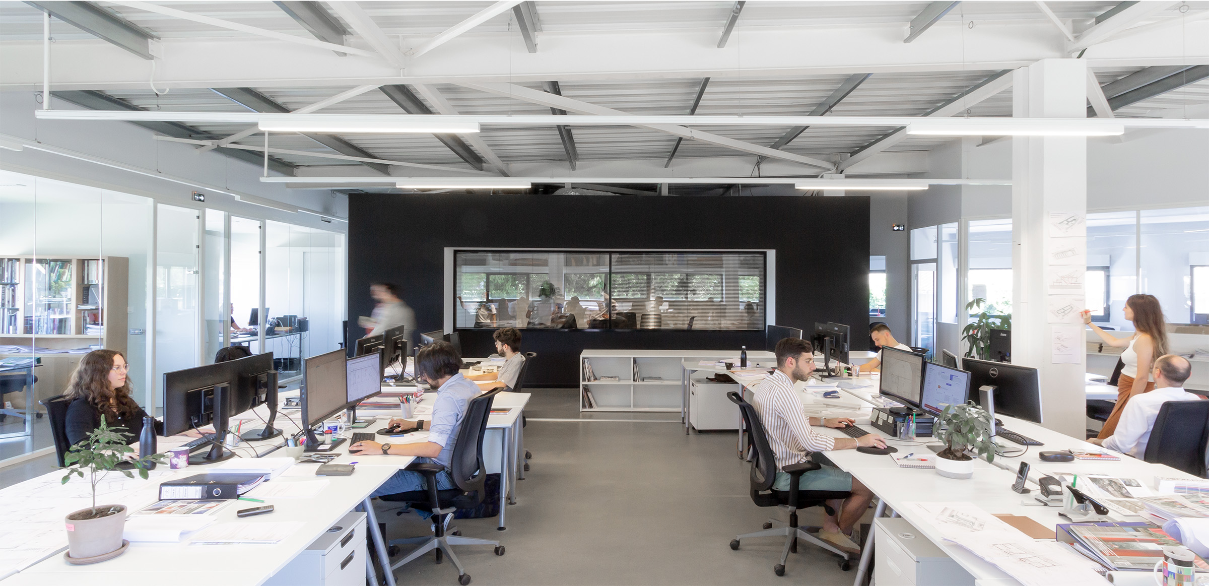 Photographie de l'ensemble du plateau vue sur la boite noire qui sert de salle de réunion entouré de tables d'architectes, lieu dominance de blanc et noir, structure métallique apparante