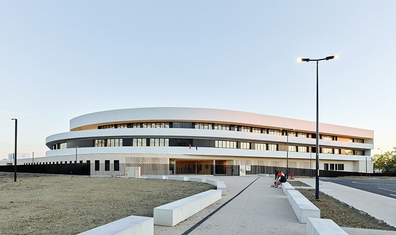 Photographie depuis l'allée centrale vers l'entrée du collège, alterance de bandeaux vitrée gris anthracite cerné d'un mur auvent blanc sur 2 niveaux en escalier