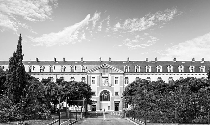 Photographie noir et blanc face à la cité administrative Reffye de Tarbes, long bâtiment sur 3 niveaux caché par une végétation dense au 1er plan