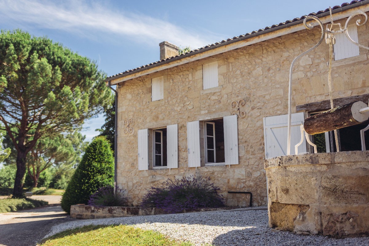 Photographie de la façade principale du château en enduit et pierre blanche, au pied de celui-ci de la lavande, devant un puits d'eau et au fond des pins