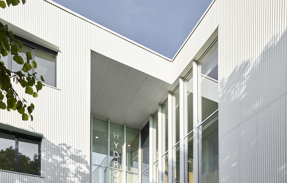 Photographie cadré du haut de l'entrée de Hydropolis à Montpellier, dans la cours entre les deux façades au bardage métallique vertical blanc, l'entrée vitrée sur deux niveaux apporte de la lumiére