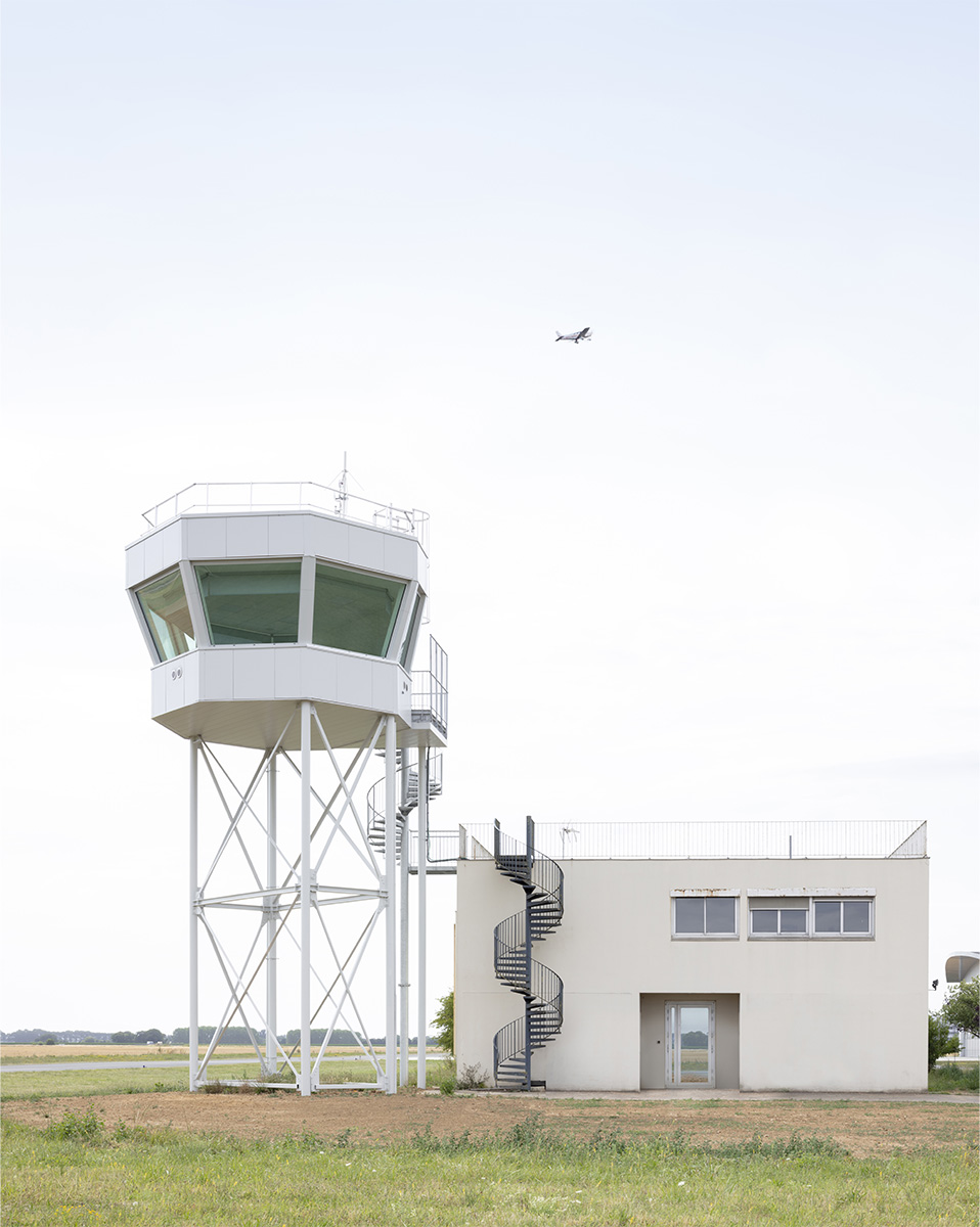 Photographie de la vigie et du bâtiment adjacent avec son accès par escalier en colimaçon par l'extèrieur, un avion en arrière plan dans le ciel