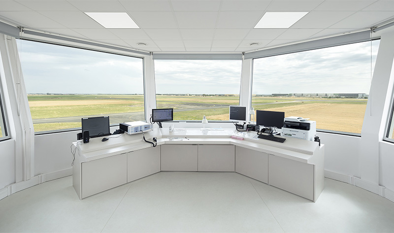 Photographie de l'intérieur de la vigie, bandeau vitré facetté en suivant la forme octogonale, mobilier blanc et écrans de surveillance, vue sur toute la piste et les champs aux alentours