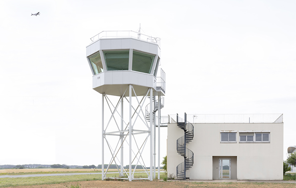 Photographie vue paysage de la vigie et du bâtiment adjacent, avec son accès par escalier en colimaçon par l'extèrieur, un avion en arrière plan dans le ciel