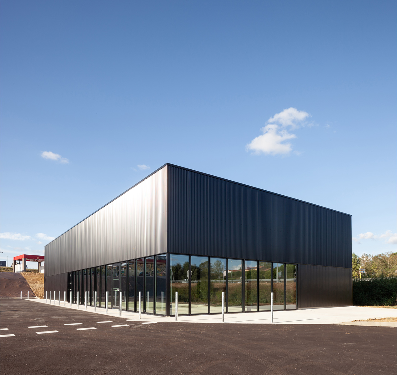 Photographie vue d'ensemble depuis le parking sur le côté du bâtiment, bardage vertical resserré gris anthracite brillant en partie haute et plus espacé en partie basse avec la partie boutique en vitrée, menuiserie gris anthracite