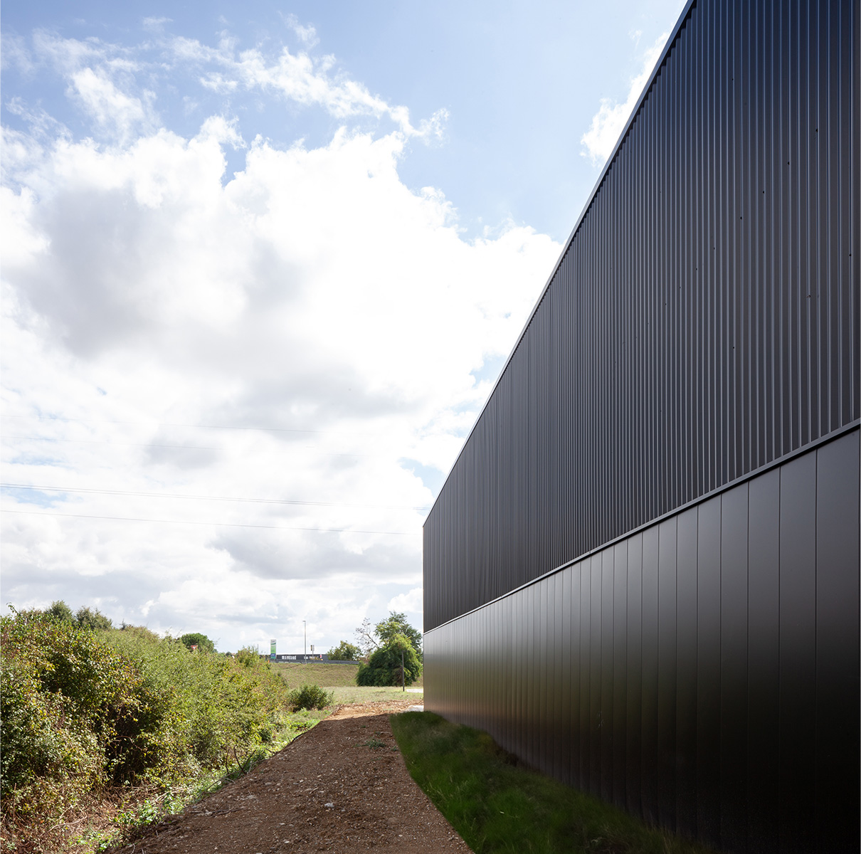 Photographie depuis l'arrière du bâtiment non visible du public, une facade grise anthracite brillante avec un bardage vertical espacé en bas et serré en haut, végétation en friche à gauche