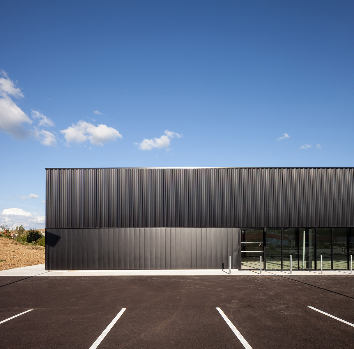 Photographie d'une partie de la longeur du bâtiment, vue depuis les places de parking en face, façade bardage vertical gris anthracite serré en hauteur et espacé en partie basse avec vitrage pour futures boutiques