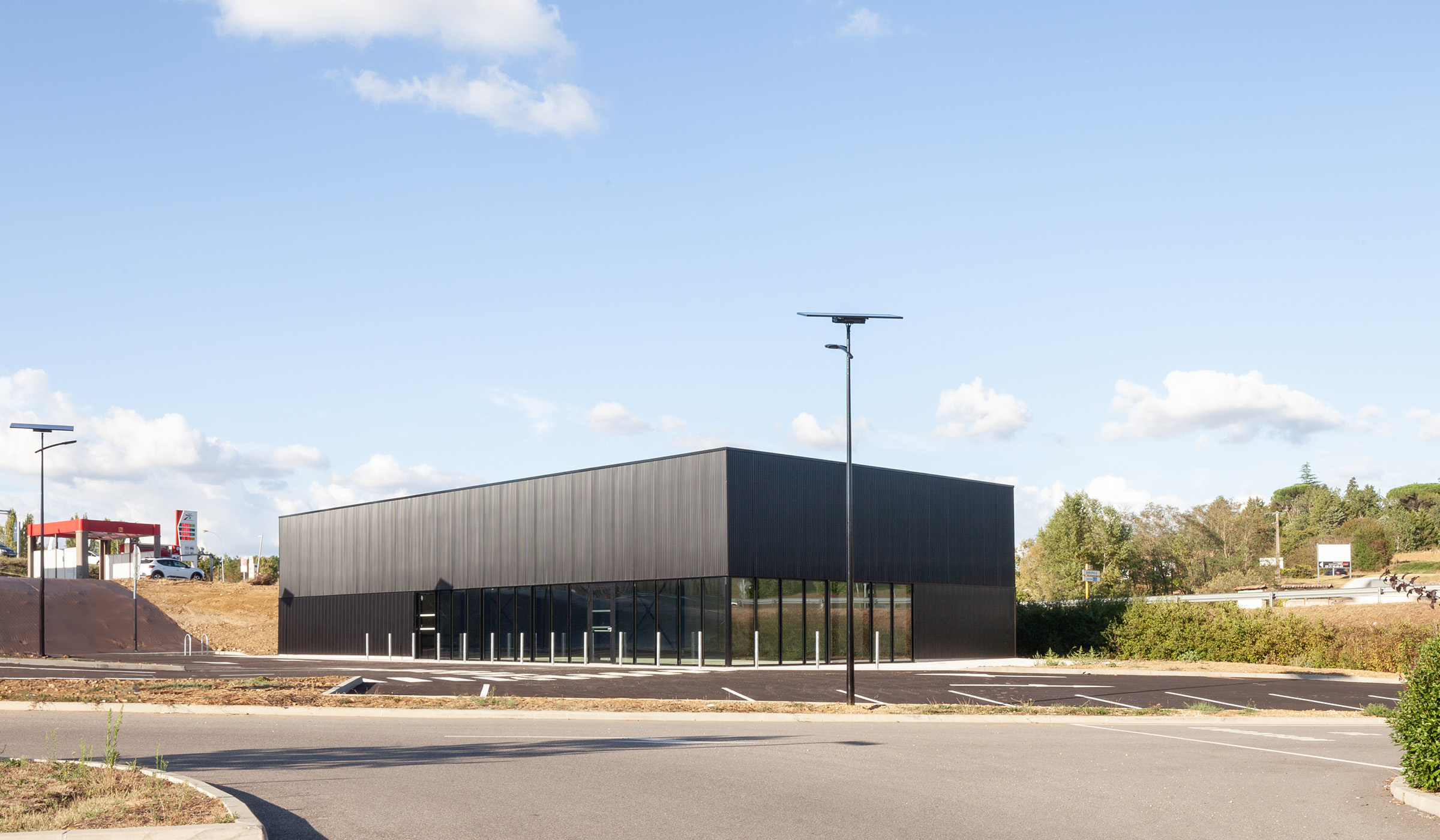 Photographie lointaine depuis un autre parking, vue d'ensemble de la coque commercial rectangle gris anthracite, bâtiment à l'ombre sur sa largeur, brillance en longeur, contexte de zone commercial à gauche et végétation à droite