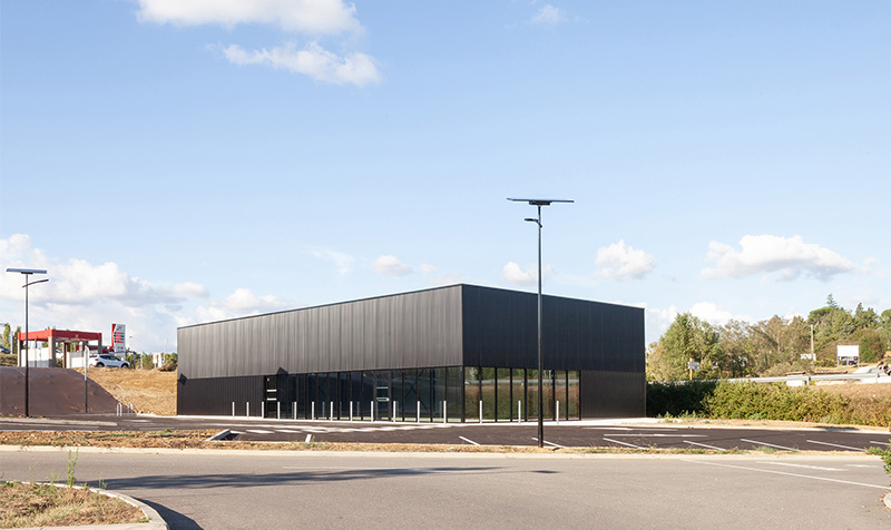 Photographie lointaine depuis un autre parking, vue d'ensemble de la coque commercial rectangle gris anthracite, bâtiment à l'ombre sur sa largeur, brillance en longeur, contexte de zone commercial à gauche et végétation à droite