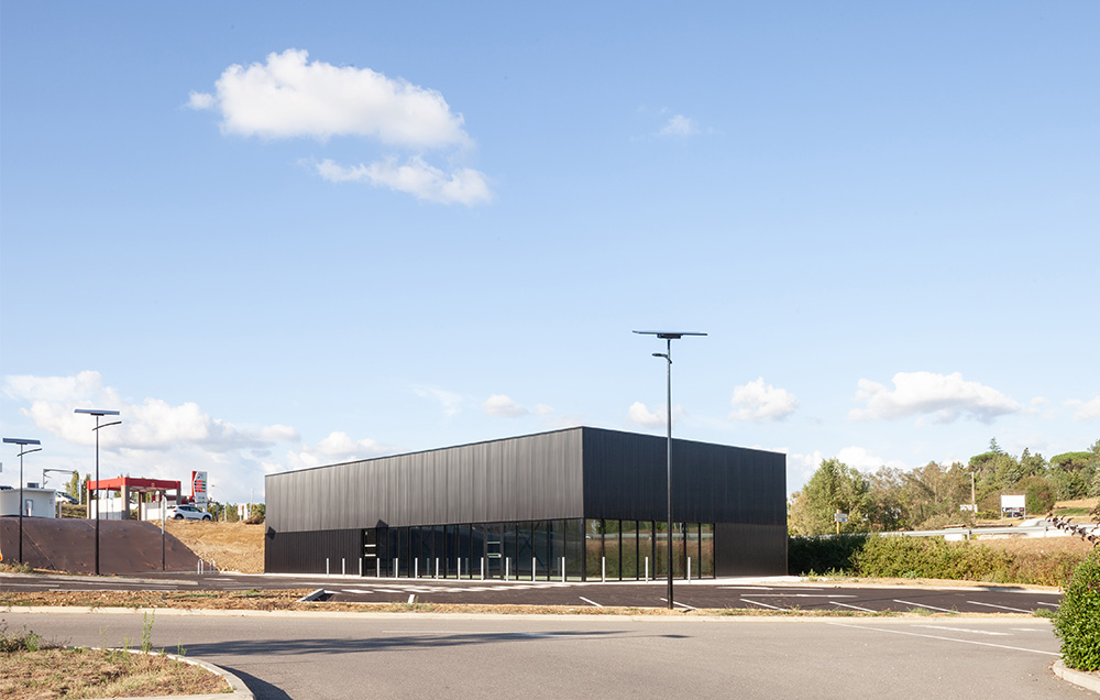 Photographie lointaine depuis un autre parking, vue d'ensemble de la coque commercial rectangle gris anthracite, bâtiment à l'ombre sur sa largeur, brillance en longeur, contexte de zone commercial à gauche et végétation à droite