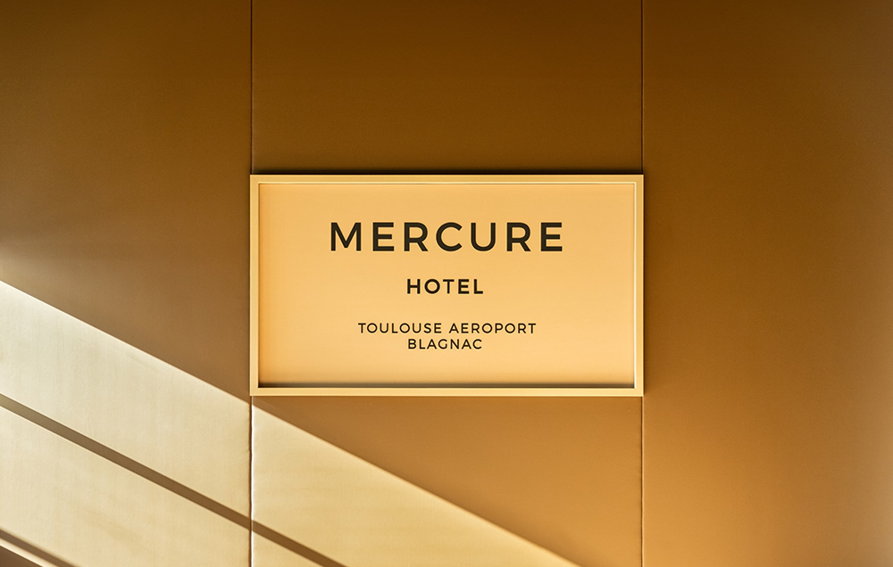 Photographie face à l'enseigne Mercure "Hotel Toulouse Aeroport Blagnac" dans un cadre jaune sur un mur ocre, l'ambiance lumineuse est dorée