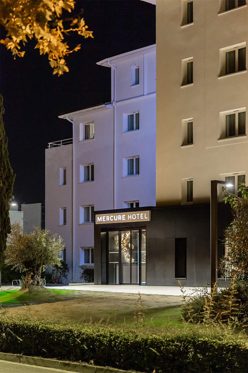 Photographie, depuis l'espace vert devant l'entrée, du bâtiment en escalier avec éclairages en façade et accueil gris anthracite avec enseigne lumineuse