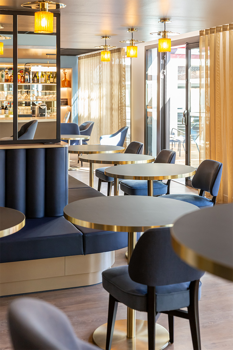 Photographie d'un espace de restauration bistrot avec tables et banquette bleu et lumières et rideaux donnent une ambiance dorée, espace lumineux avec la terrasse