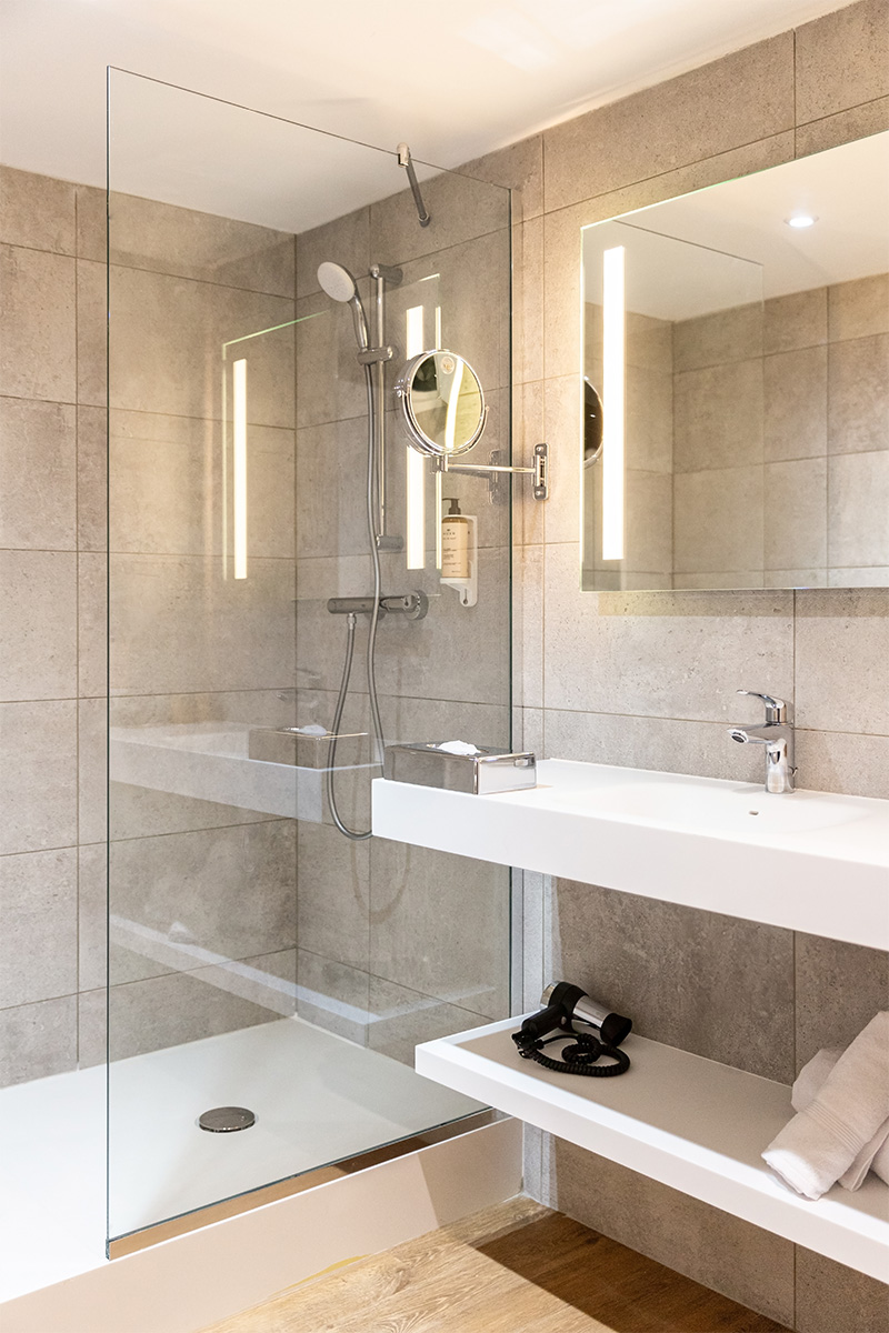 Photographie d'une salle d'eau avec douche cabine, sol en bois, mobiliers et plafond blanc, mur en grands carreaux de couleur pierre