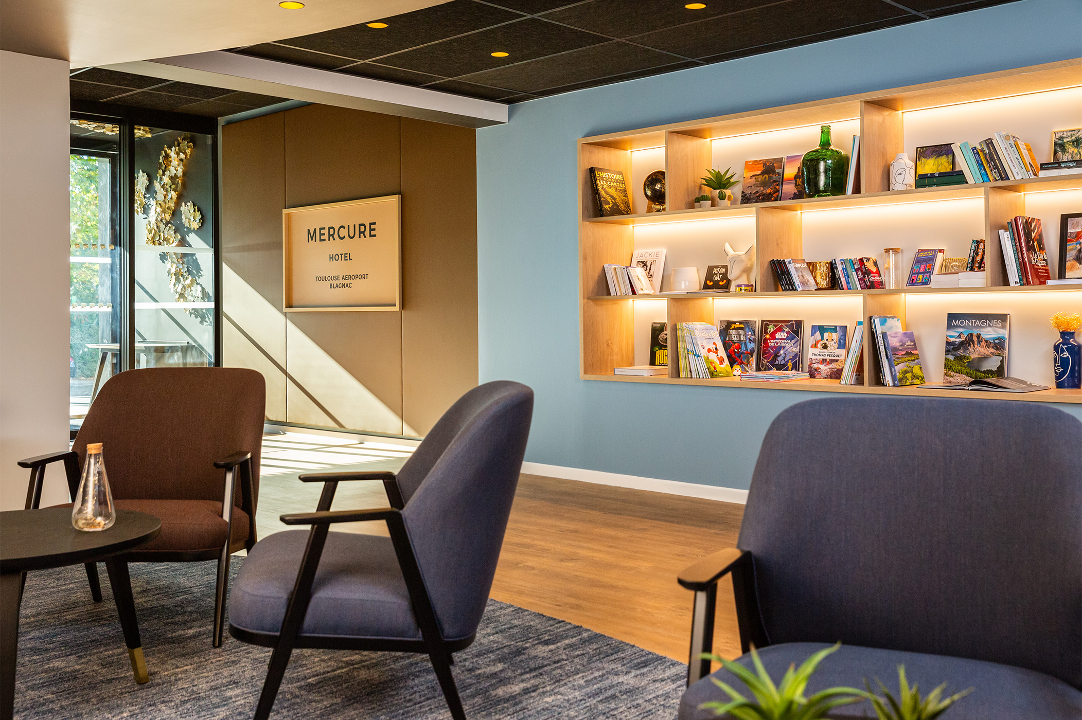 Photographie du hall d'entrée depuis l'espace détente avec les tables et fauteuils, vue sur une librairie lumineuse, sol en bois et murs bleu clair