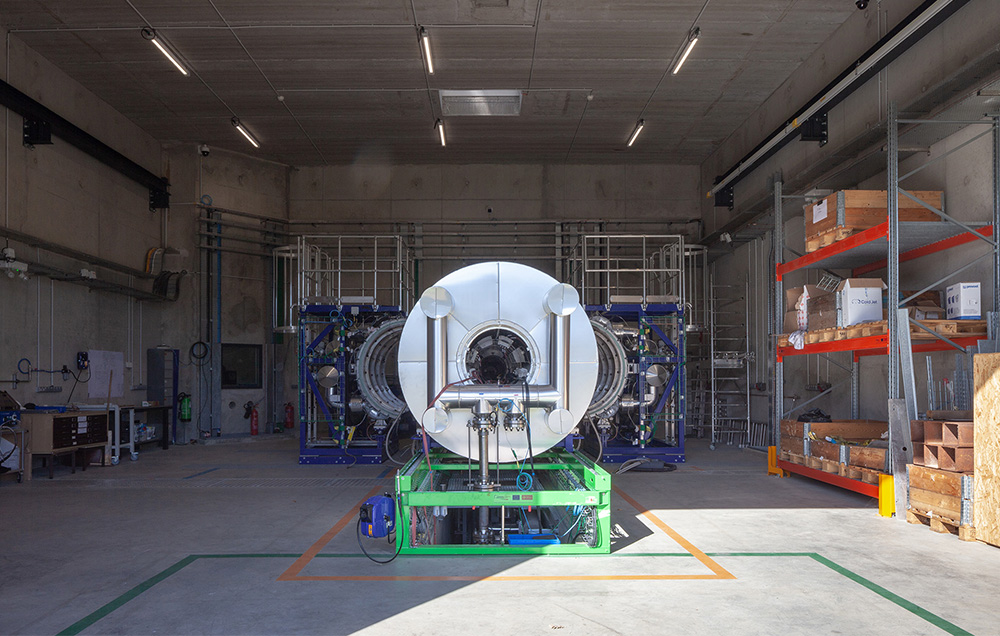 Photographie depuis l'entrée d'un garage d'expérimentation en béton de l'ONERA - Toulouse, structure en métal avec des tuyaux et au 1er plan face à un cylindre métallique avec pleins de capteurs, marquage rectangulaire au sol pour délimiter les périmètres des process