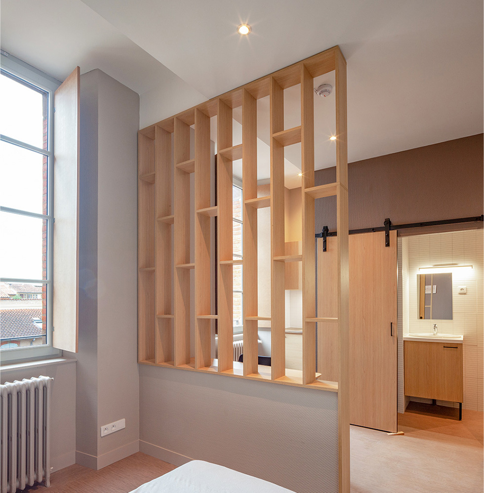 Photographie d'une chambre rénovée vue sur les toits avec deux fenêtres, au milieu une étagère cadres en bois tout hauteur, mobiliers et sol teinte bois, mur gris et marron clair, plafond blanc