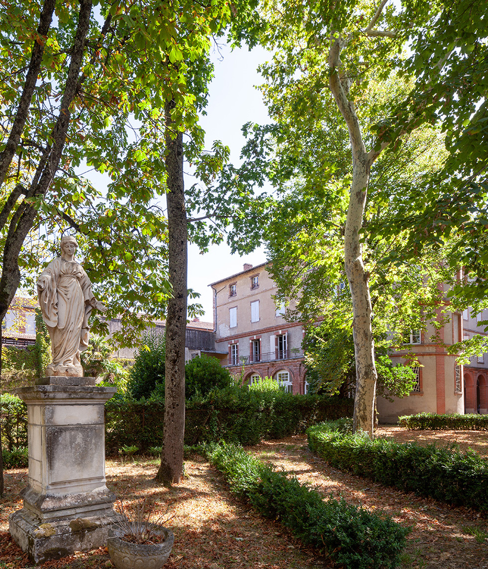 Photographie depuis le jardin sous l'ombre des grands arbres verdoyant, vue sur le diocèse au loin derrière la végétation