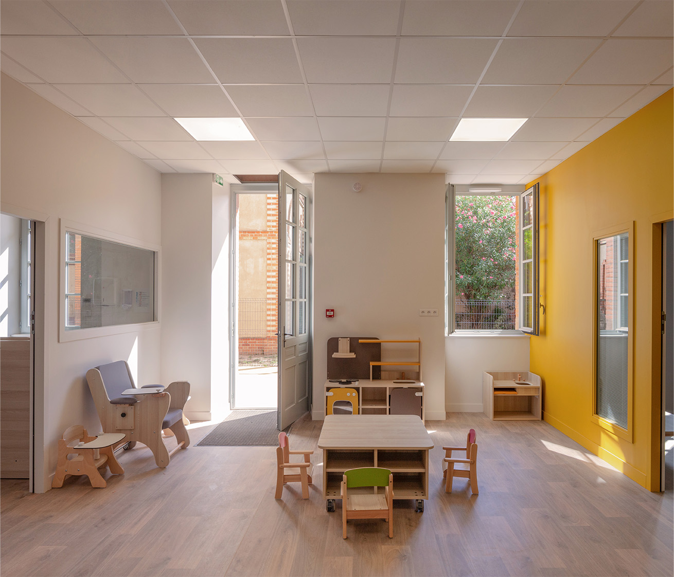 Photographie d'une salle de jeux pour les enfants, une porte d'entrée et fenêtre donnent sur une cour, de petits mobiliers en bois attendent d'être découvert, sol en bois et mur blanc avec un pan en jaune orangé