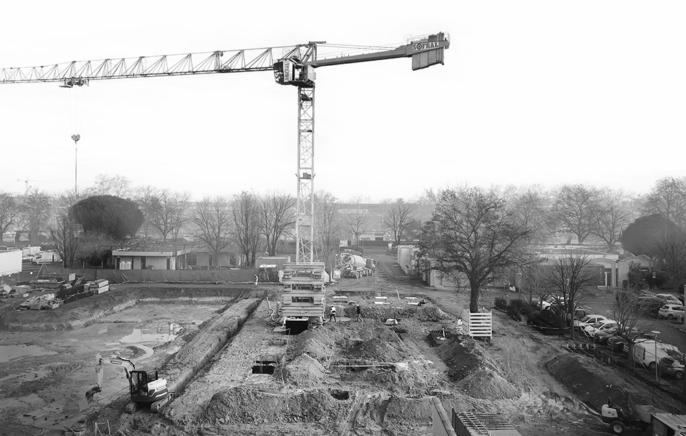 Photographie noir et blanc en hauteur du début du chantier avec le sol creusé, aplani avec des monticules de terres et une grue au milieu, arbres d'hiver aux alentours sans leurs feuillages