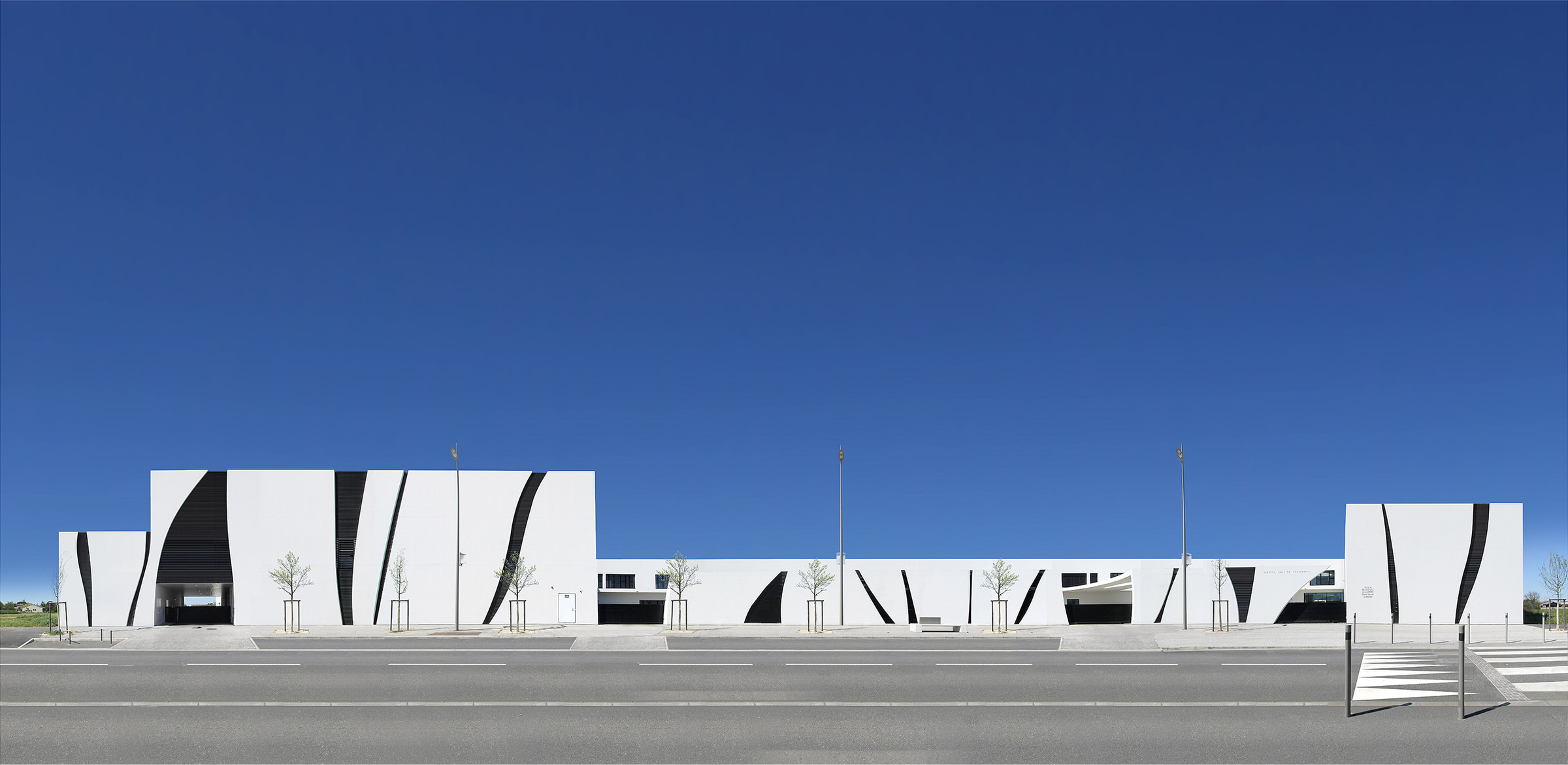 Photographie de la façade de l'école noir et blanc depuis le trottoir d'en face, bâtiment au différentes hauteurs rectangulaires blanches soulignés d'arabesques noires, le ciel est bleu profond